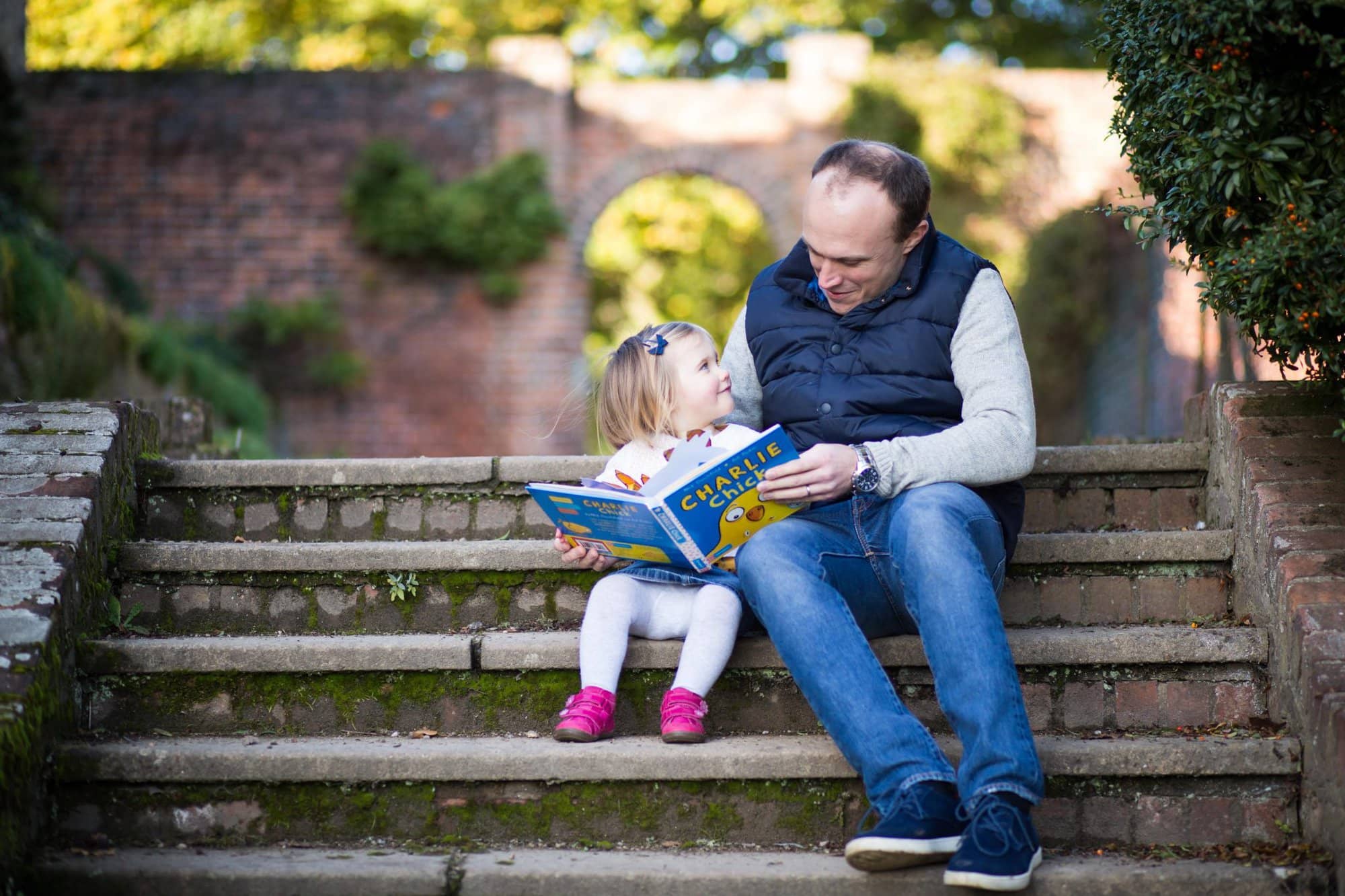 Photo of Dad and young daughter sitting on steps reading book, daughter looking lovingly into Dad's eyes in Beckenham, a lovely natural family moment captured
