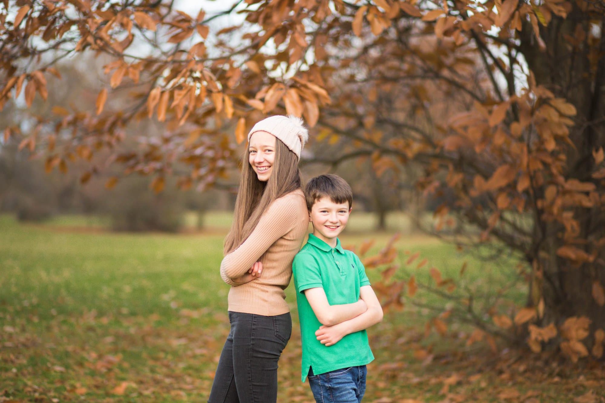 Brother and sister leaning against each other under a tree of orange Autumn leaves, smiling and natural portrait of a family having fun in Beckenham Place Park