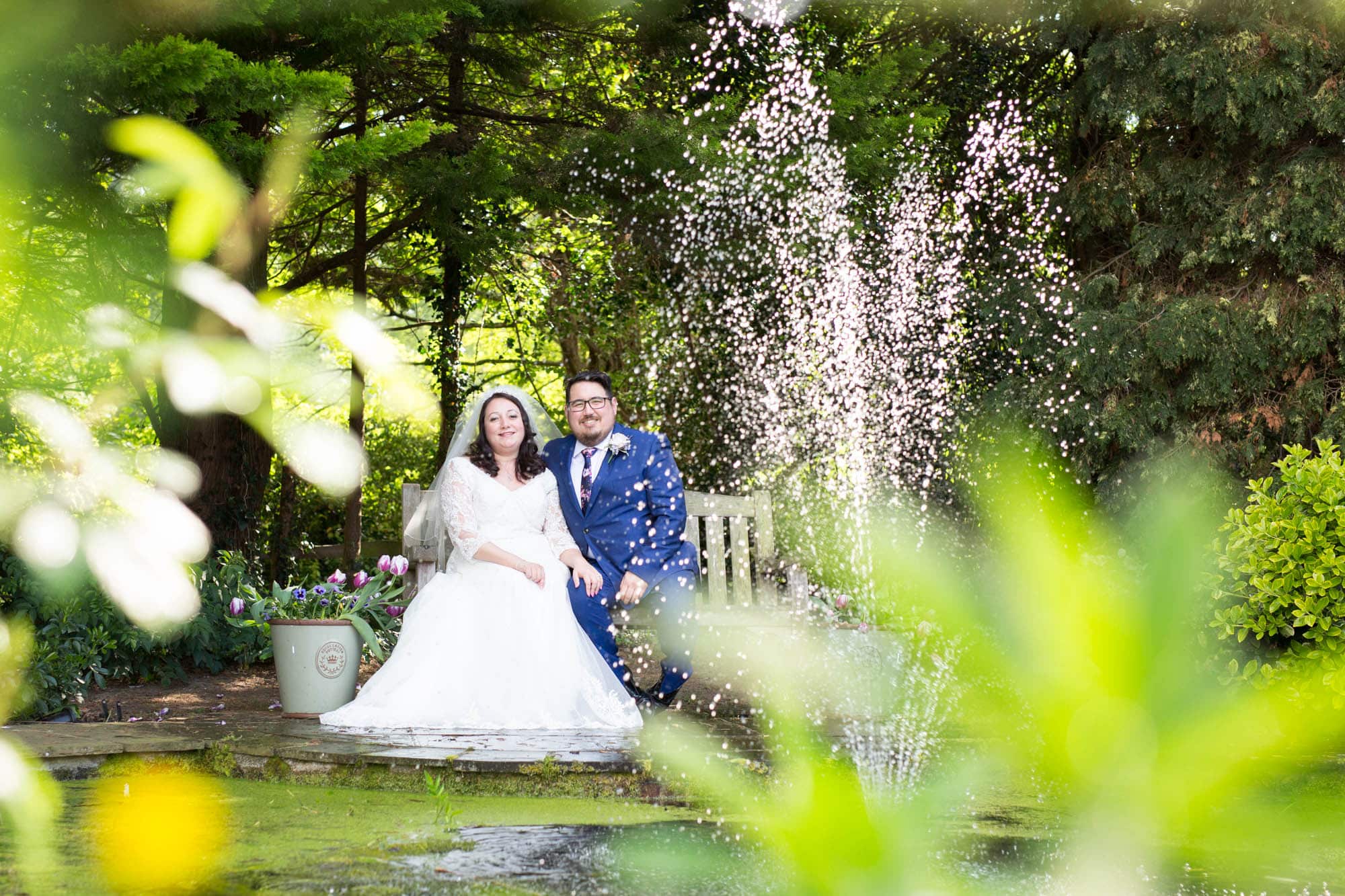 Smiling wedding couple sat on bench by waterfall and surrounded by greenery at Oaks Farm Weddings venue in Croydon