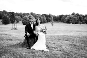 Wedding couple sitting in Beckenham place park on log smiling at Beckenham photographer in black and white photo