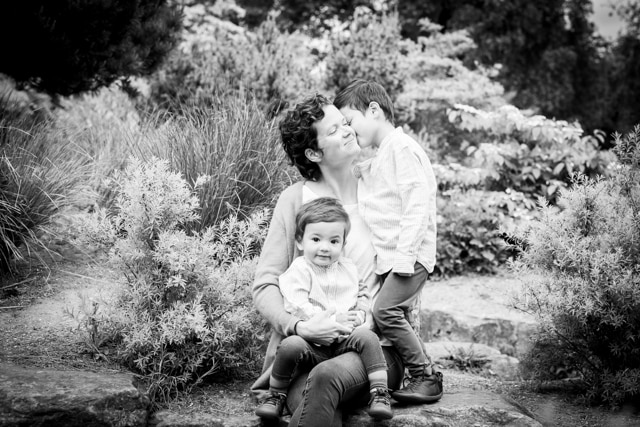 Mum being kissed by eldest son outside in park while younger son sits on lap being cuddled and smiling in family photoshoot