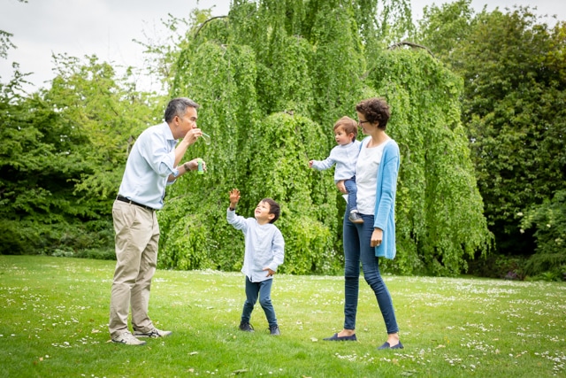 Dad blowing bubbles while children try to catch them with mum holding youngest on her hip in outdoor family Bromley photoshoot