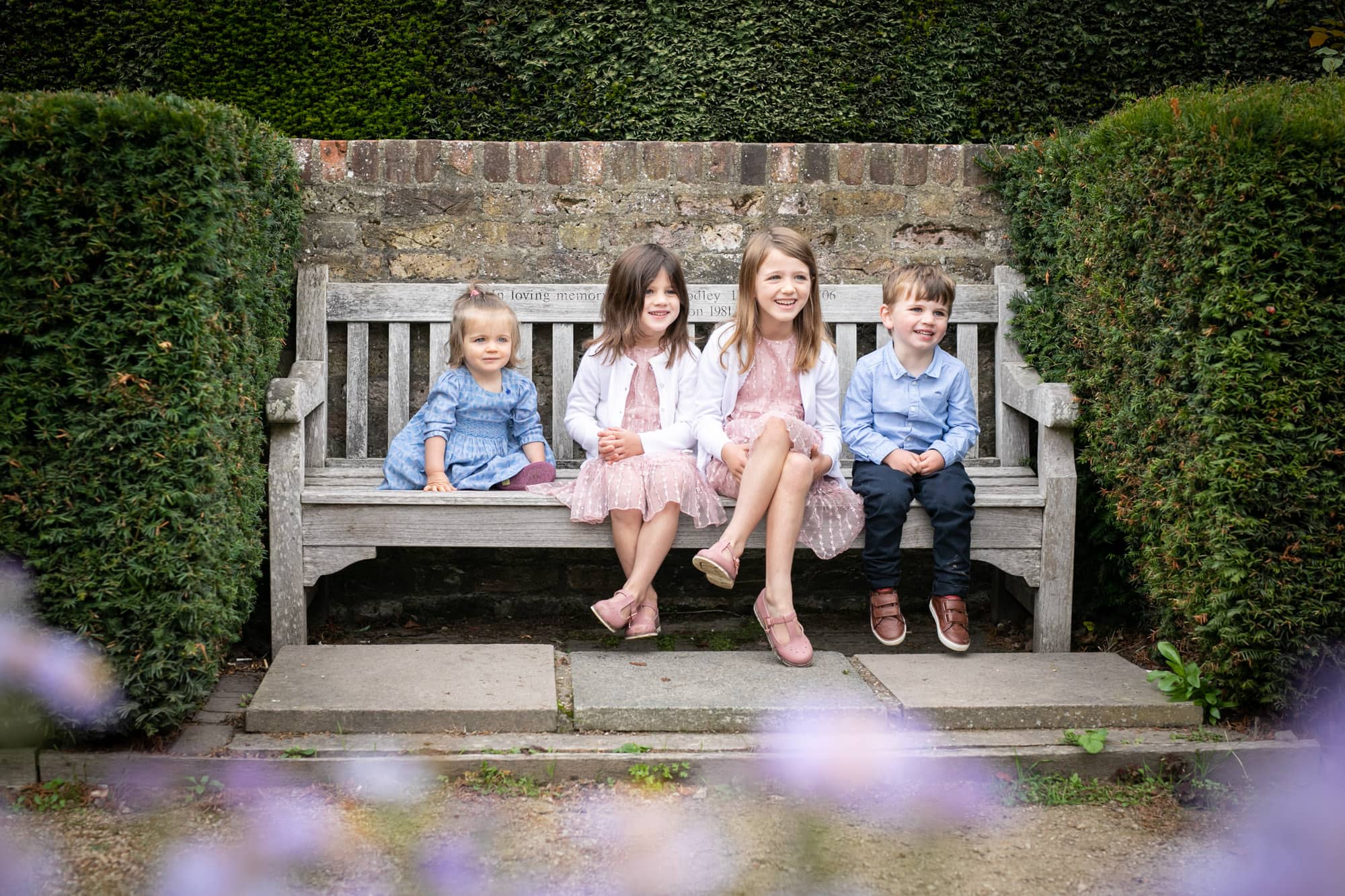 Siblings sat on bench and looking away from camera with flowers in foreground taken by Bromley Photographer
