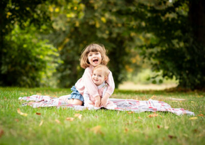 Sisters cuddling and laughing sat on blanket outside on grass in photoshoot in Bromley