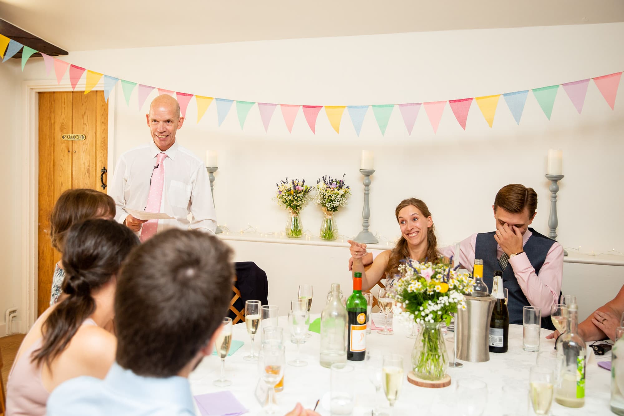 Father of bride giving wedding speech with groom hiding his face and bride pointing at bridesmaid in wedding photo by Bromley wedding photographer at Oaks Farm Weddings
